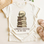 Ivory t-shirt with a skull on books graphic and 'Currently Haunted by my TBR' text, placed on a light surface.