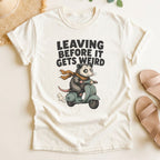 Ivory t-shirt with a graphic of a possum on a scooter and text 'Leaving Before It Gets Weird' on a light background.