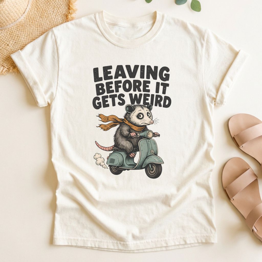Ivory t-shirt with a graphic of a possum on a scooter and text 'Leaving Before It Gets Weird' on a light background.